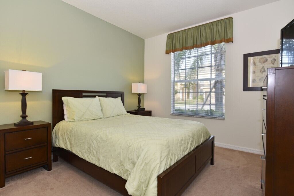 A bedroom with a bed, two bedside tables with lamps, a window with blinds, and a framed picture on the wall.