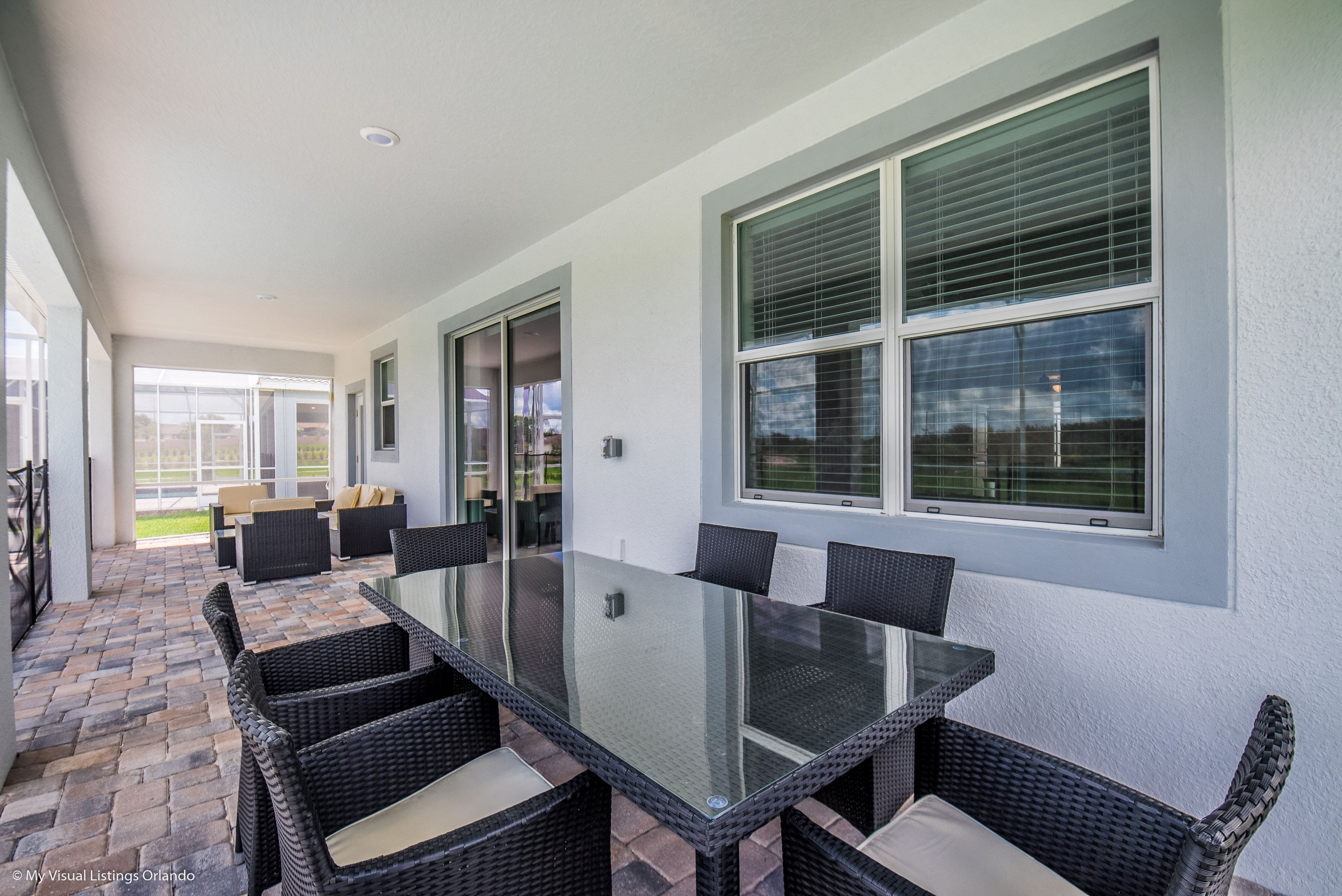 A covered patio area with a glass-top table and wicker chairs, large windows with blinds, and a view of the outdoors.