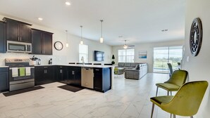 A modern kitchen with dark cabinetry, a central island, and a dining area with yellow chairs. - 9 Bedroom Champions Gate Resort (Davenport)