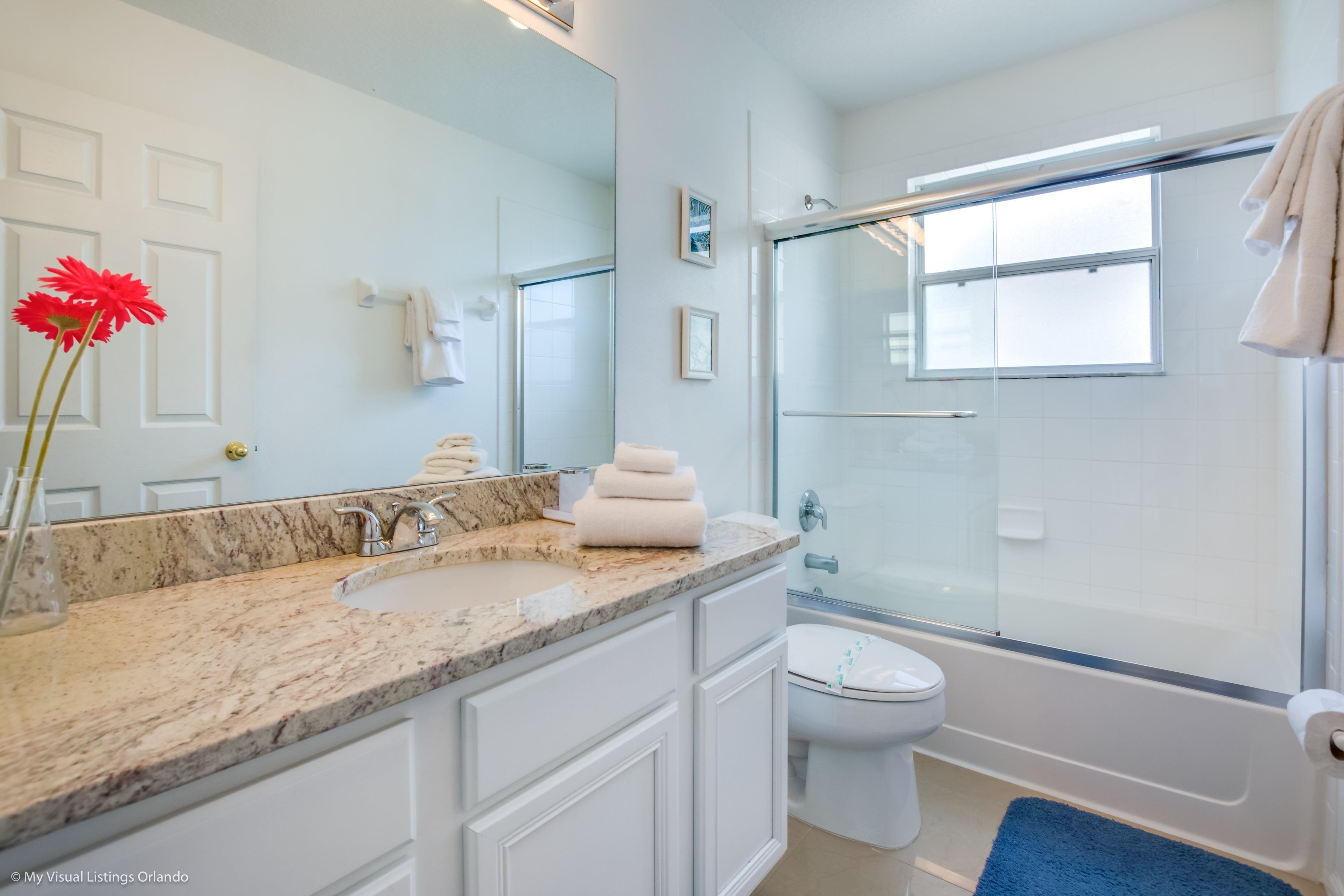 A modern bathroom with a marble sink, white cabinets, a glass shower enclosure, and a blue bath mat.