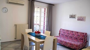 Un coin repas comprenant une table, des chaises, un saladier de fruits, un radiateur, un climatiseur, une fenêtre avec des rideaux et un canapé.