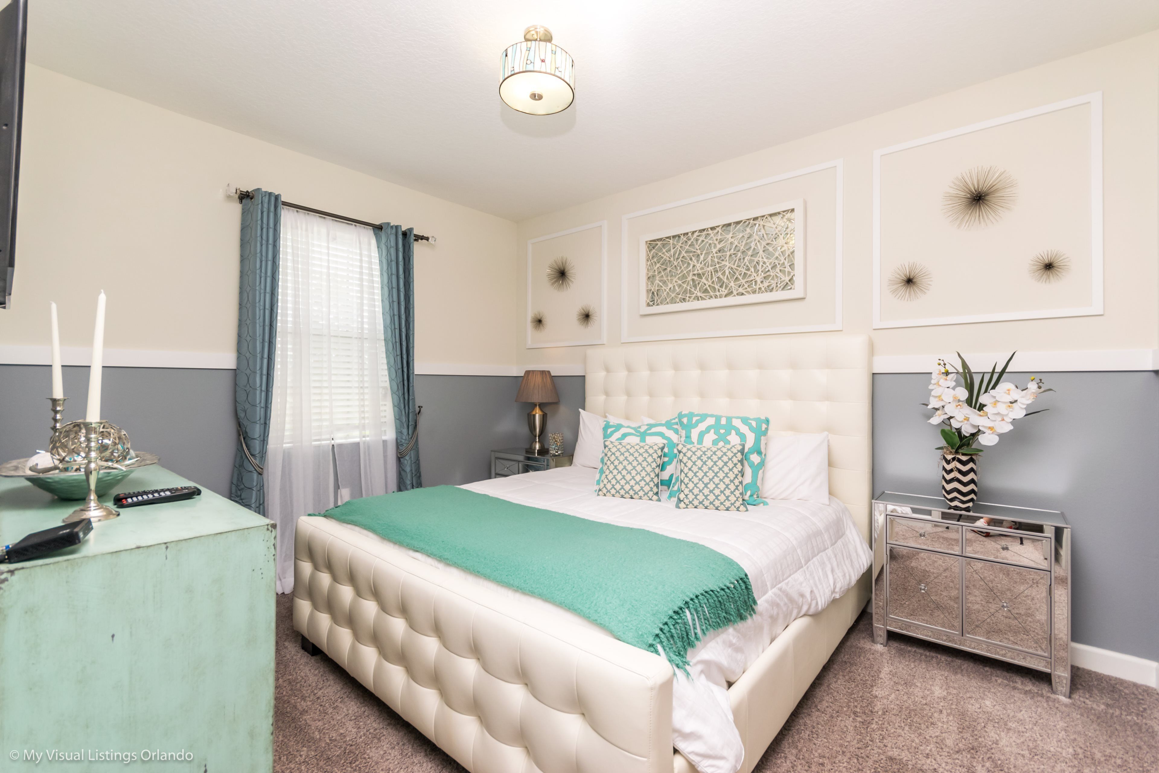 Une chambre à coucher avec une tête de lit capitonnée, un plaid vert, une table de chevet avec une lampe et des œuvres d’art décoratives accrochées au mur.