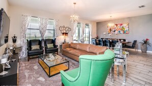 A spacious living room with a brown leather sofa, a green armchair, and a coffee table. There is a fireplace with decorative items and a chandelier. The room has large windows with curtains and a dining area in the background. - 1314bw00 (Davenport)