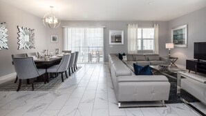 A modern living room with a white sofa, dining area with a table and chairs, and a large window with blinds. - 8 Bdrm 5 Bath Villa (Davenport)
