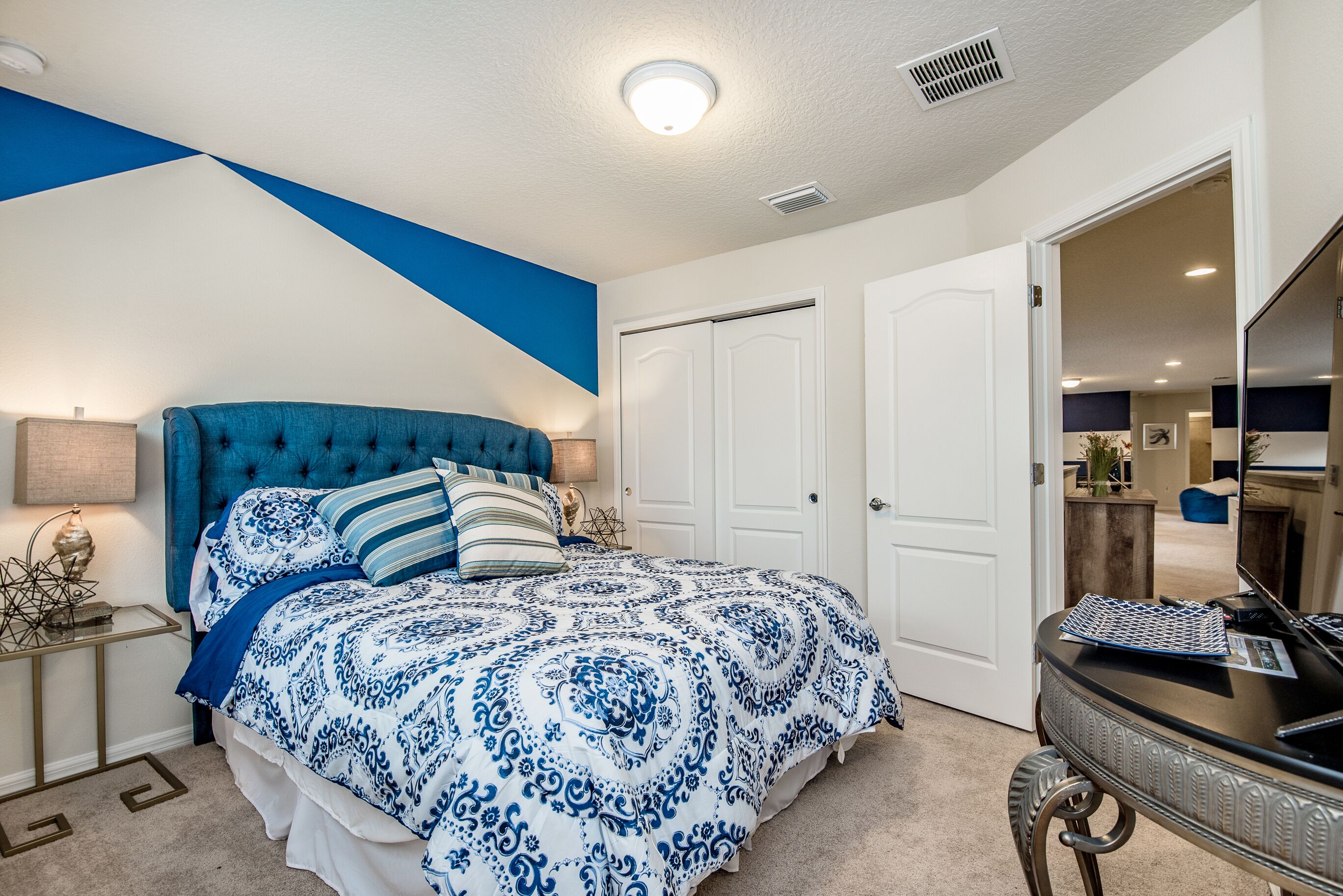 A bedroom with a tufted headboard, a large bed with a blue and white patterned bedspread, a bedside table with a lamp, and a dresser with a mirror.