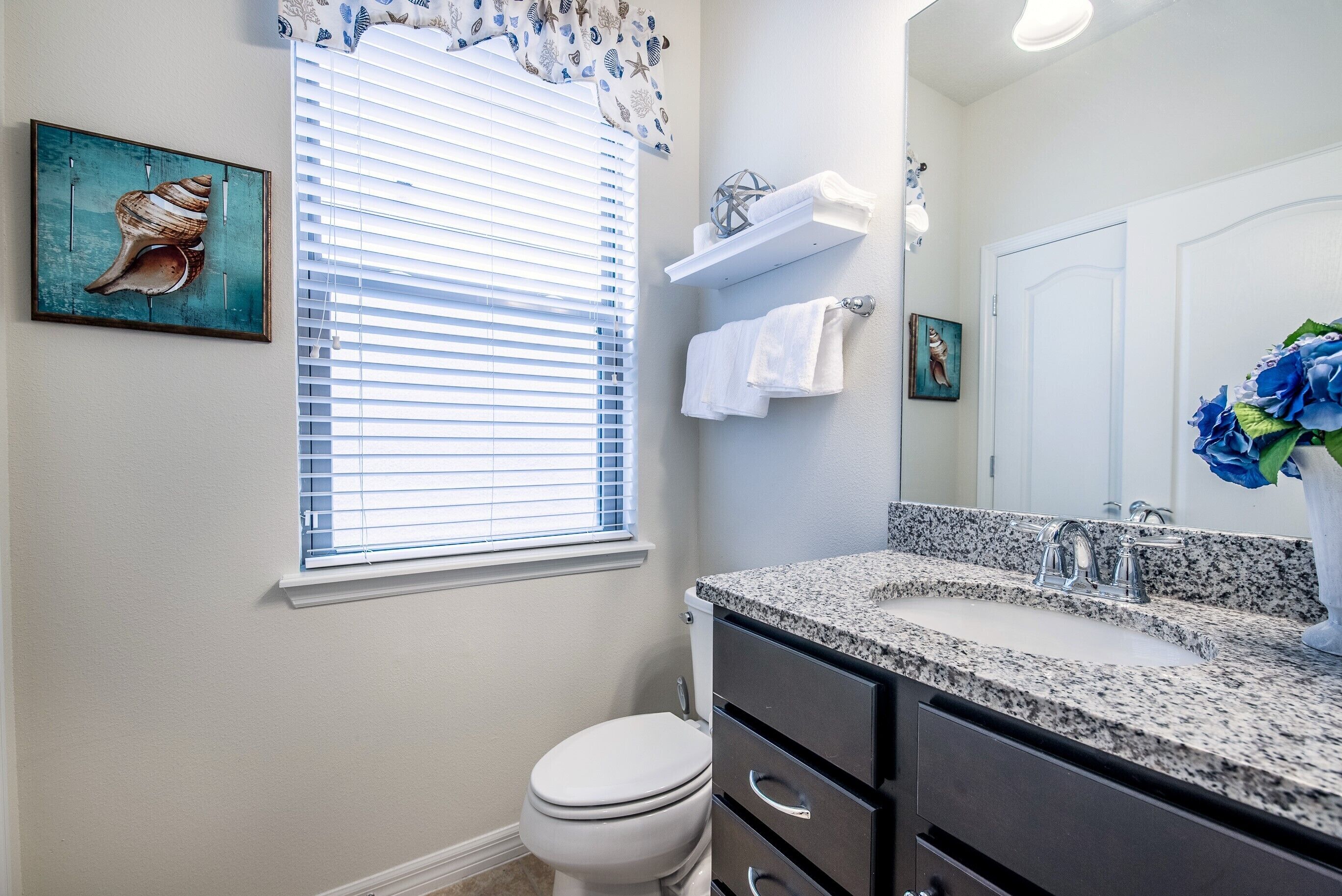A bathroom with a toilet, sink, and vanity. There is a window with blinds, a towel rack, and a framed picture on the wall.