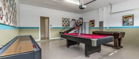 A pool table with a red felt surface, a foosball table, and a shuffleboard table in a room with a tiled floor and a white ceiling.