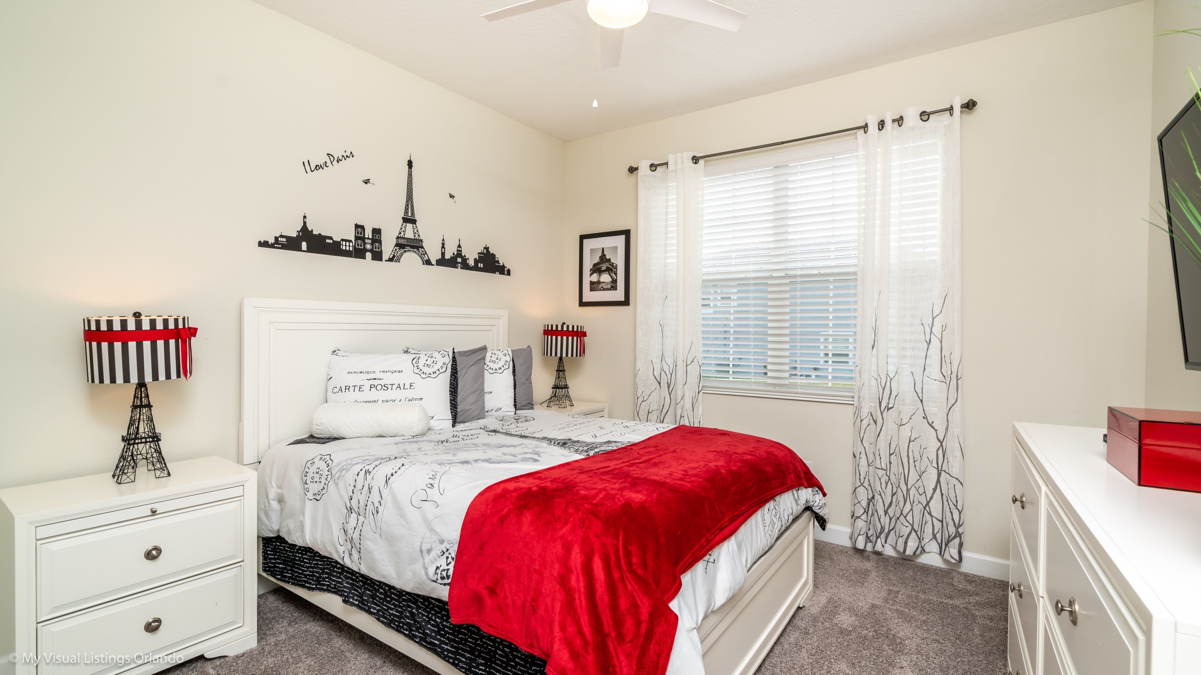 Un dormitorio con cama, mesitas de noche, cómoda, ventana con persianas, ventilador de techo y decoración en la pared, incluyendo una vista urbana y la silueta de la Torre Eiffel.