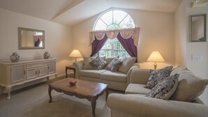 Un salon chaleureux avec un plafond haut, une grande fenêtre avec des rideaux, un ensemble de canapés, une table basse en bois et un buffet agrémenté d’objets décoratifs.