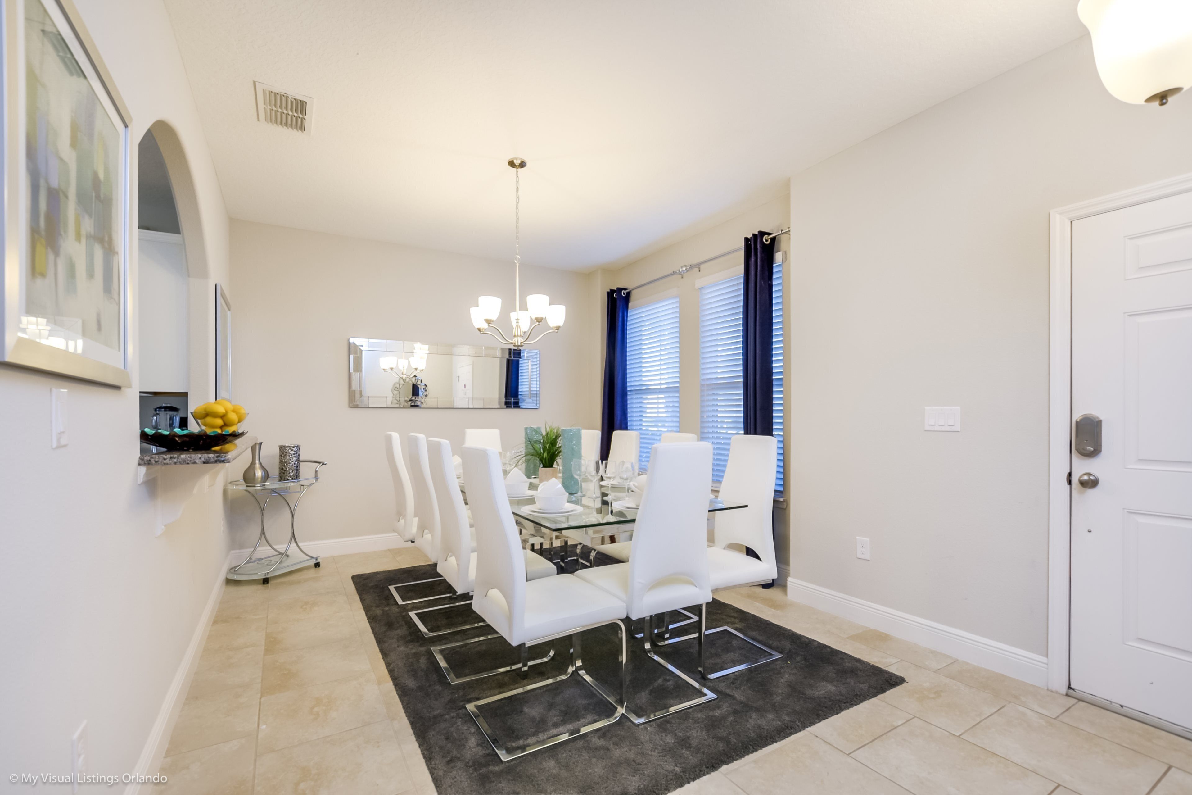A dining area with a glass table, white chairs, a chandelier, and a black rug.