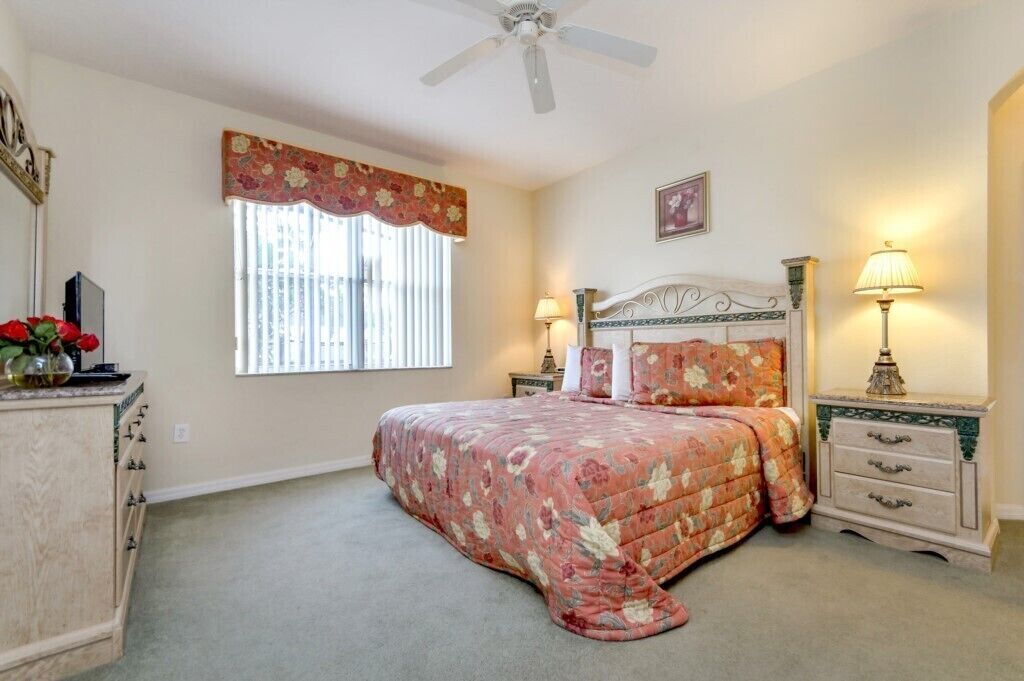 A bedroom with a bed, two bedside tables with lamps, a dresser, a ceiling fan, a window with blinds, and a floral-patterned bedspread.