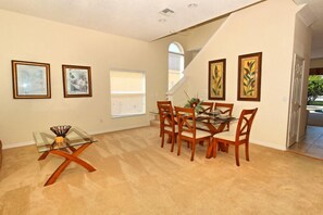 A dining area with a glass table and wooden chairs, framed artwork on the walls, and a staircase leading to another level.