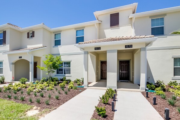 A two-story residential building with numbered doors, a tiled roof, and a landscaped front yard.