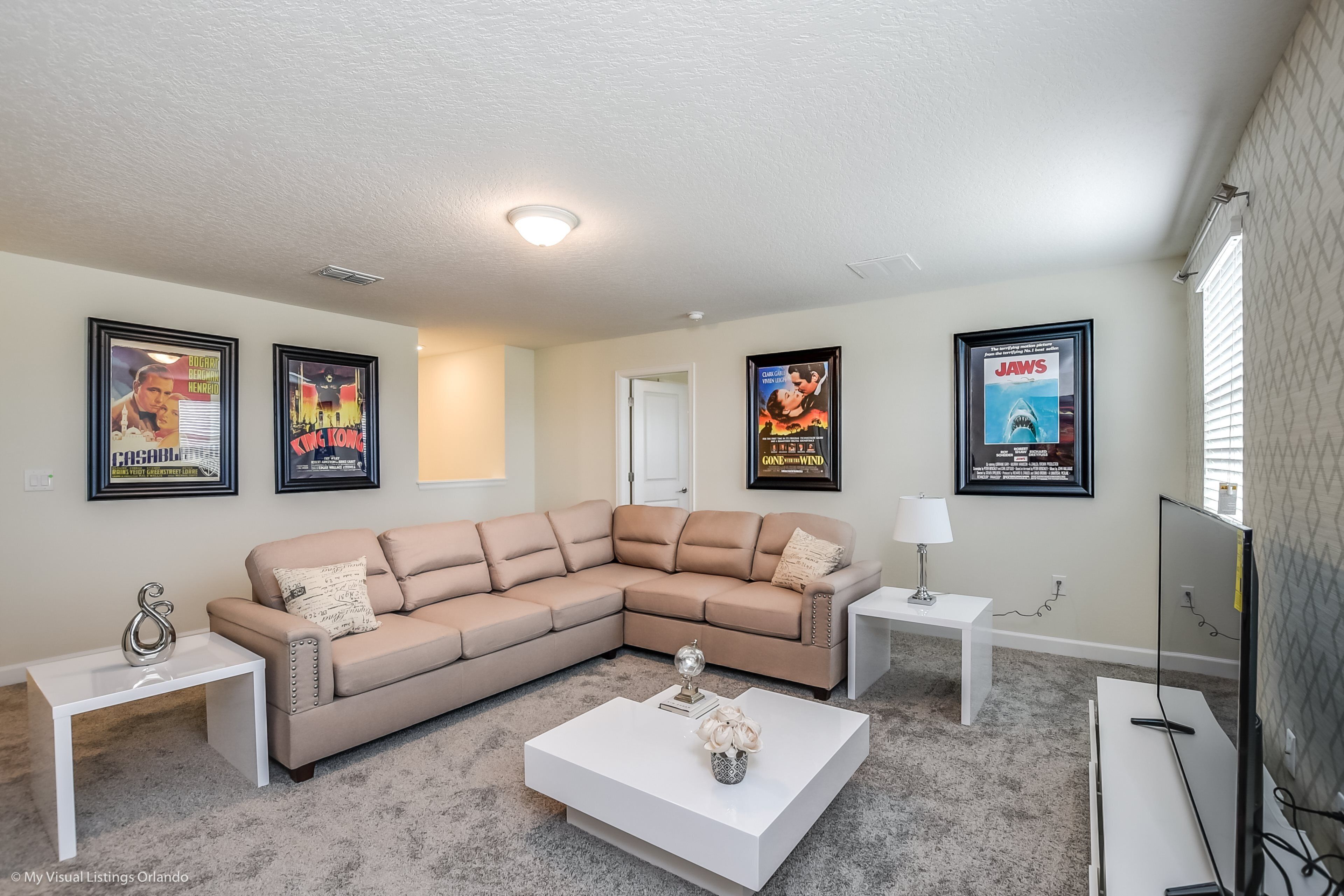 A living room with a beige sectional sofa, a white coffee table, and a TV on a stand. There are framed movie posters on the wall and a small white table with a lamp.