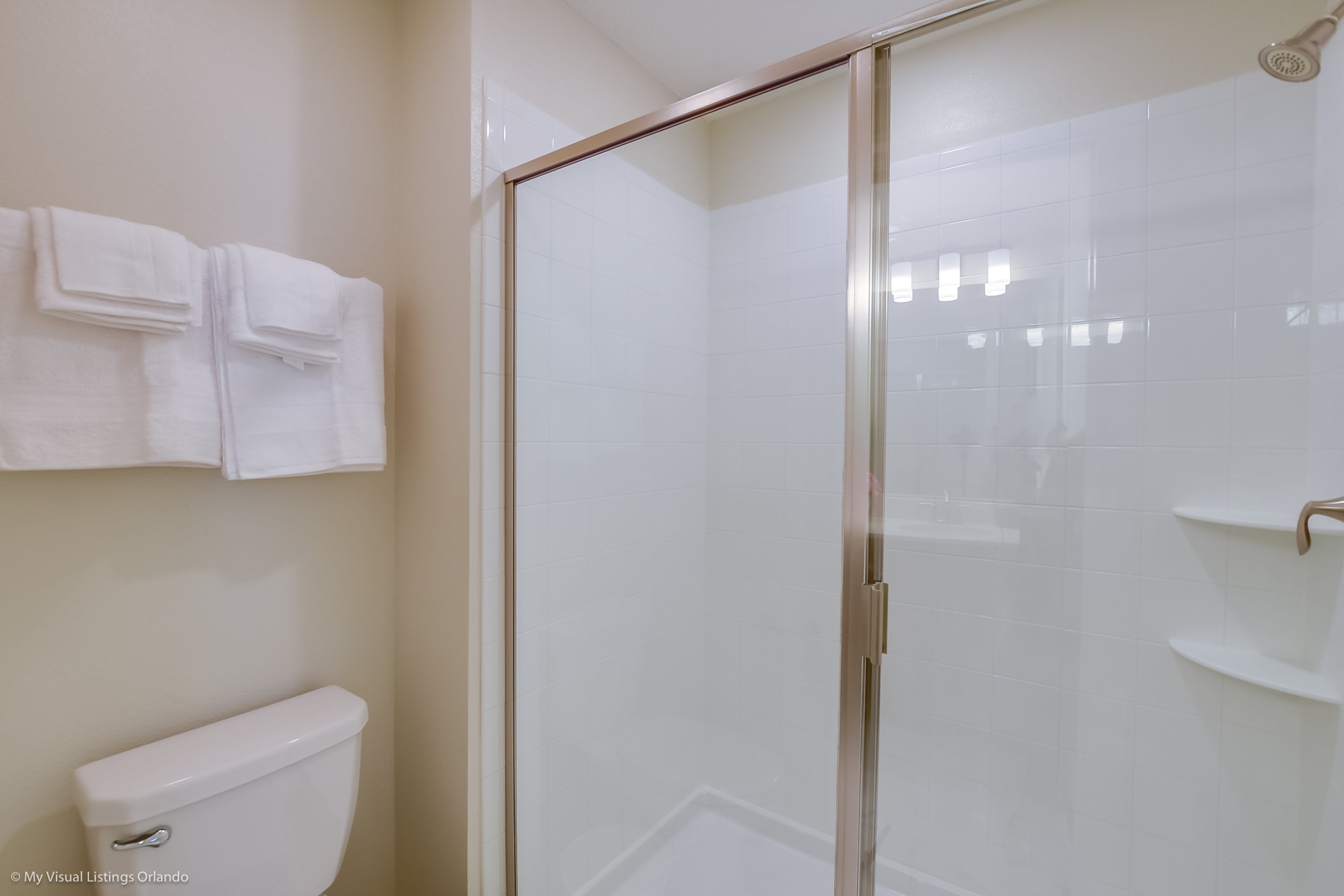 A bathroom with a glass-enclosed shower, white tiles, a toilet, and a towel rack with folded towels.