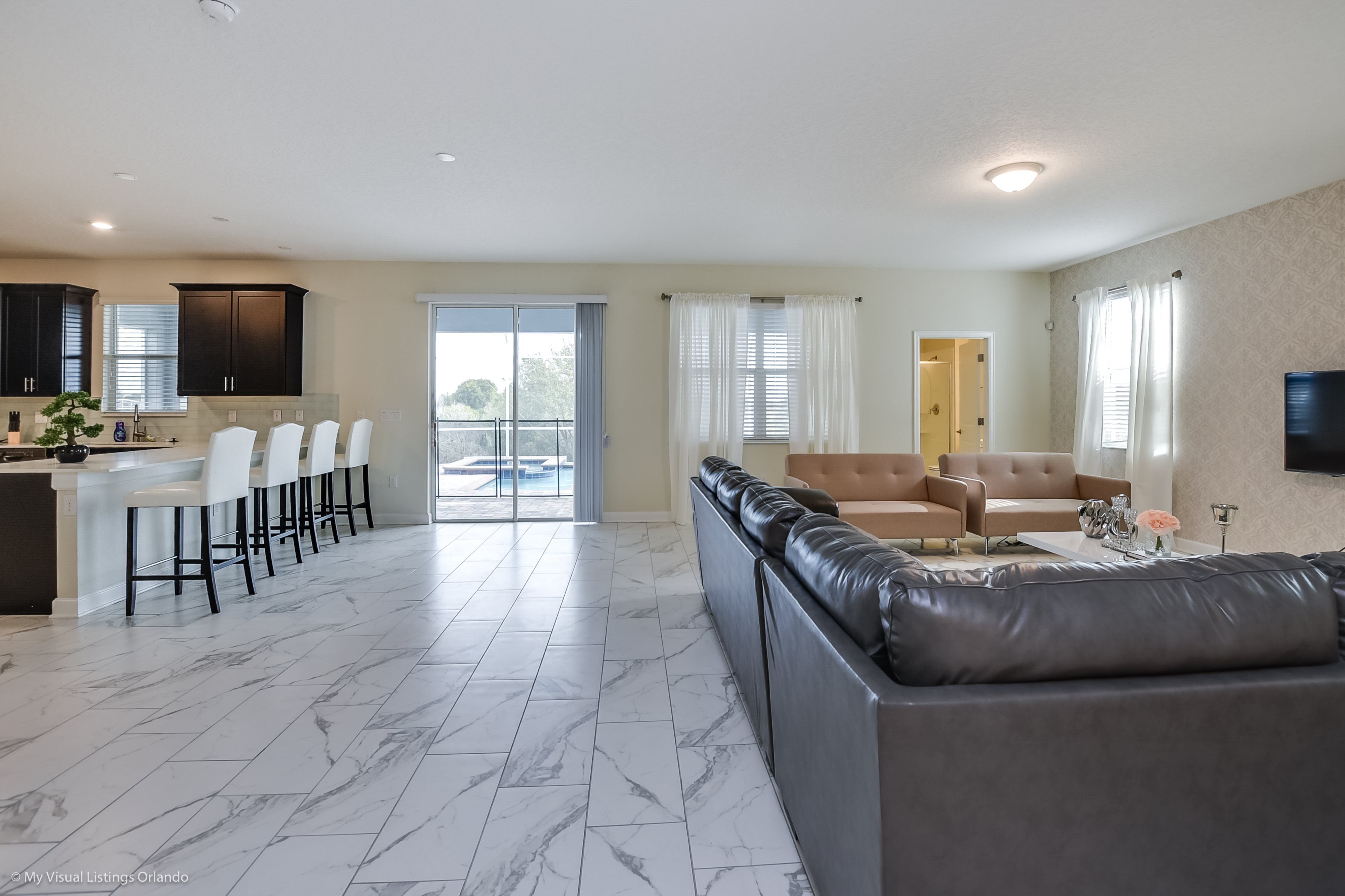 Un salon moderne avec un canapé en cuir gris, un coin repas avec des chaises blanches et une cuisine avec des meubles sombres.