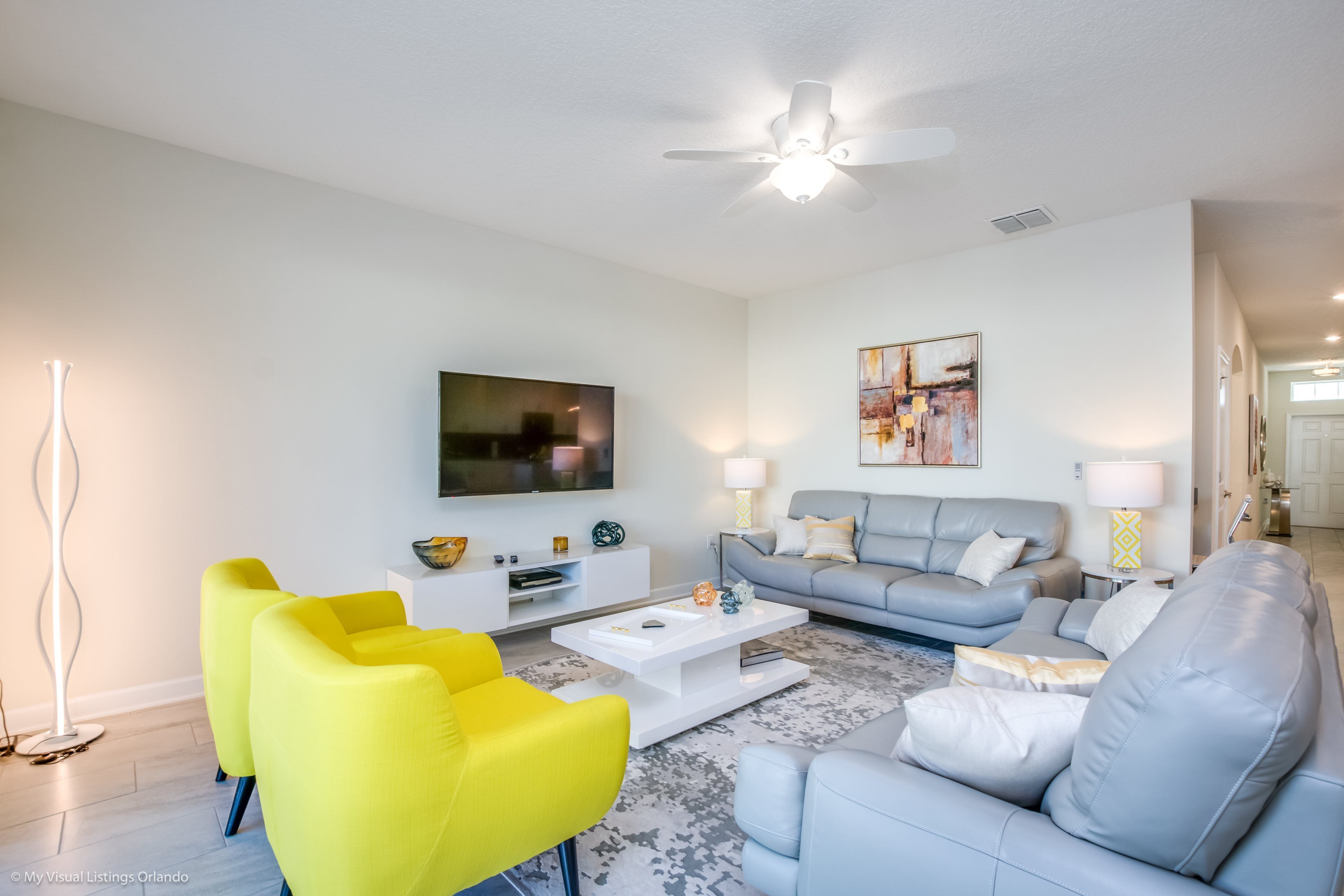 Una sala de estar moderna con un televisor de pantalla plana, un sofá gris, sillones amarillos y una mesa de centro blanca.