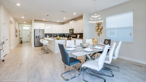 A modern kitchen with white cabinets, a central island, and a dining area with white chairs and a glass-top table. - High End Champions Gate 6 Bdrm (Davenport)