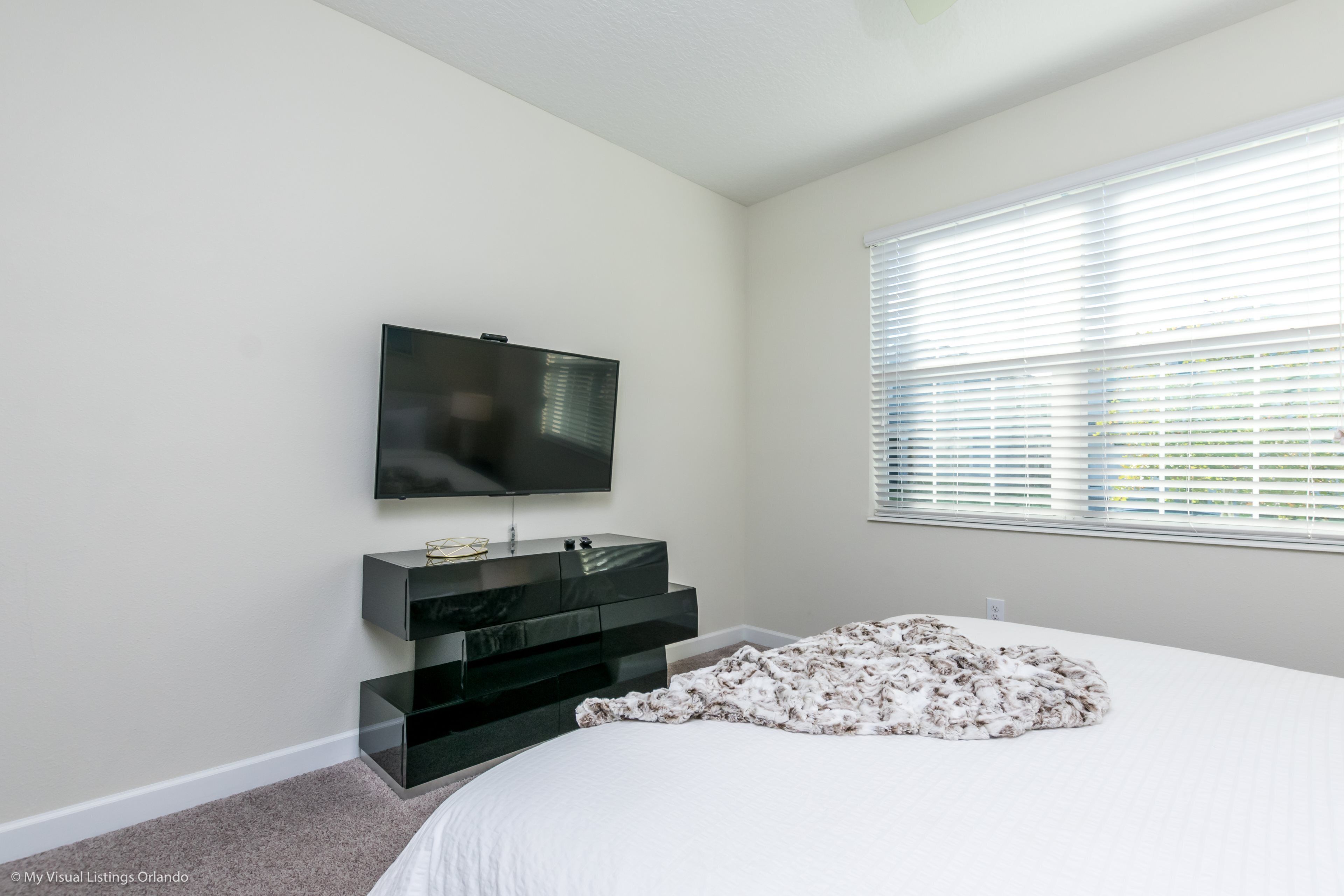 Une chambre à coucher avec un lit, un téléviseur fixé au mur, une console noire et une fenêtre avec des stores.