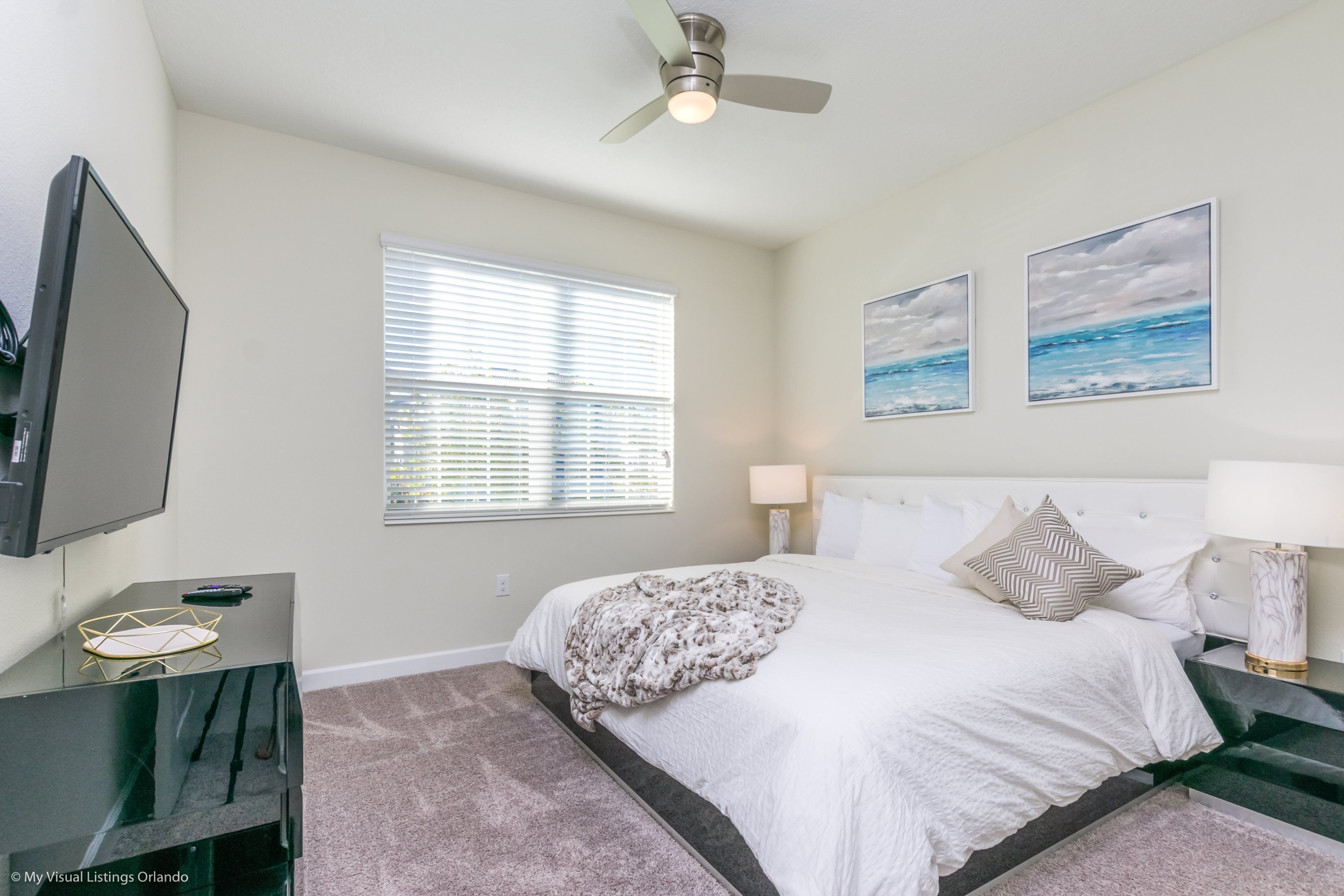 A bedroom with a bed, a TV, a ceiling fan, a window with blinds, and two wall-mounted paintings.