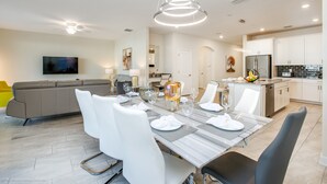 A modern kitchen with white cabinets, a stainless steel refrigerator, and a dining area with a marble table and white chairs. - High End Champions Gate 6 Bdrm (Davenport)