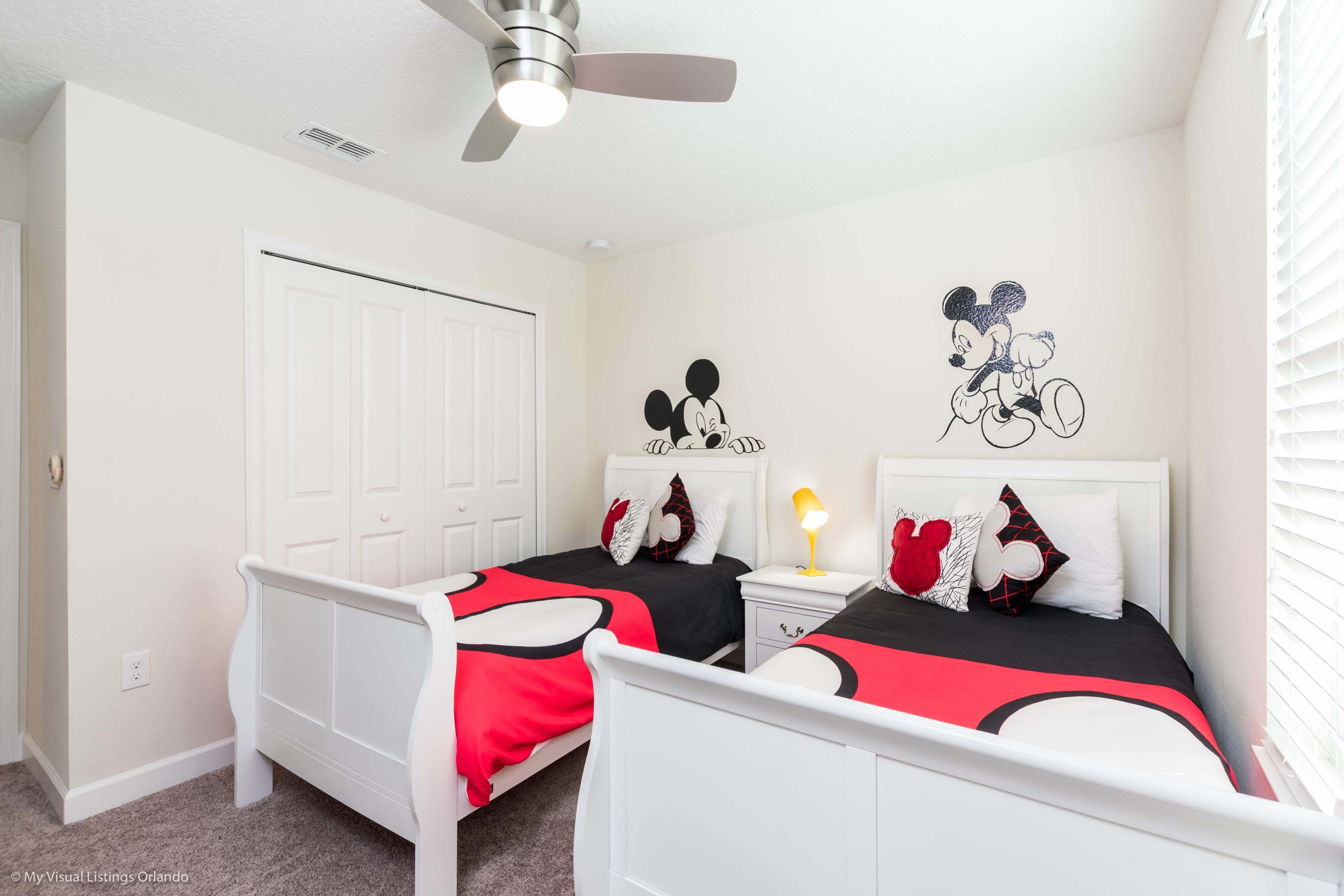 A bedroom with two beds, white headboards, a ceiling fan, and a Mickey Mouse wall decal.