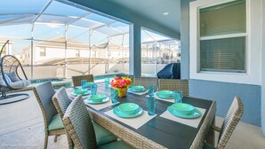 An outdoor dining area with a table set for four, surrounded by wicker chairs, a ceiling fan, and a view of buildings in the background. - High End Champions Gate 6 Bdrm (Davenport)