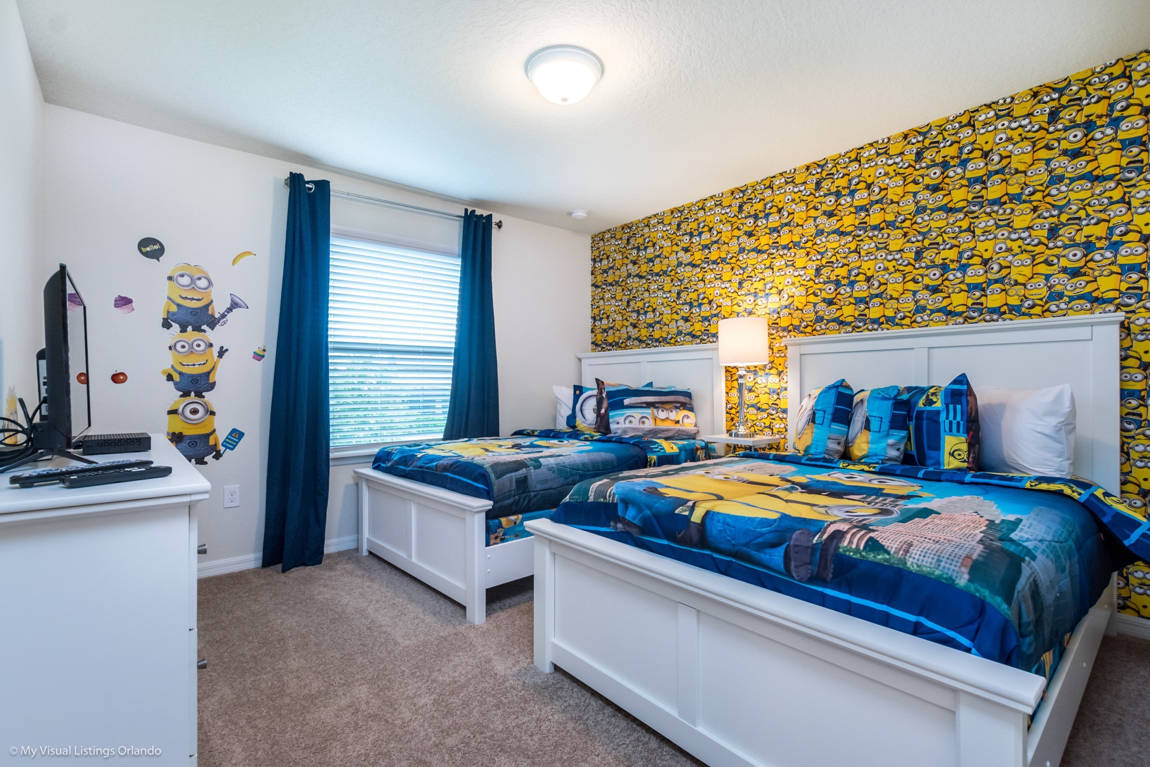 A bedroom with two beds, a desk, and a TV. The walls are decorated with a Minion-themed wallpaper.