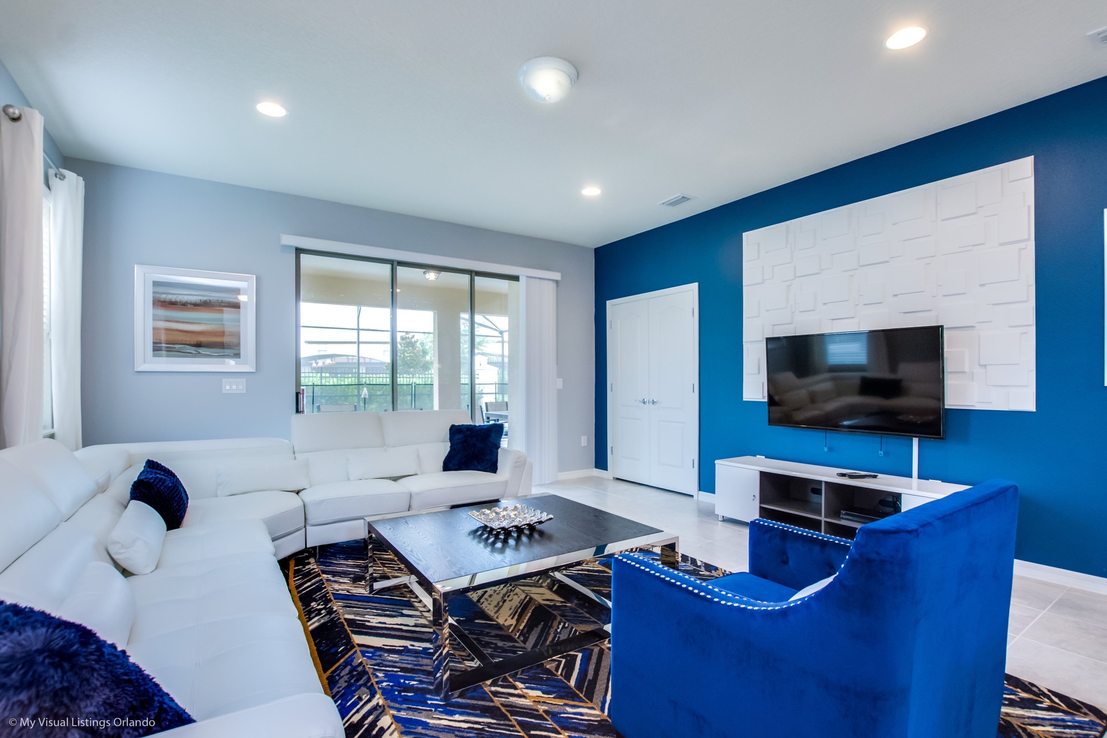 A modern living room with a white sectional sofa, a blue armchair, a coffee table, and a flat-screen TV mounted on a blue accent wall.