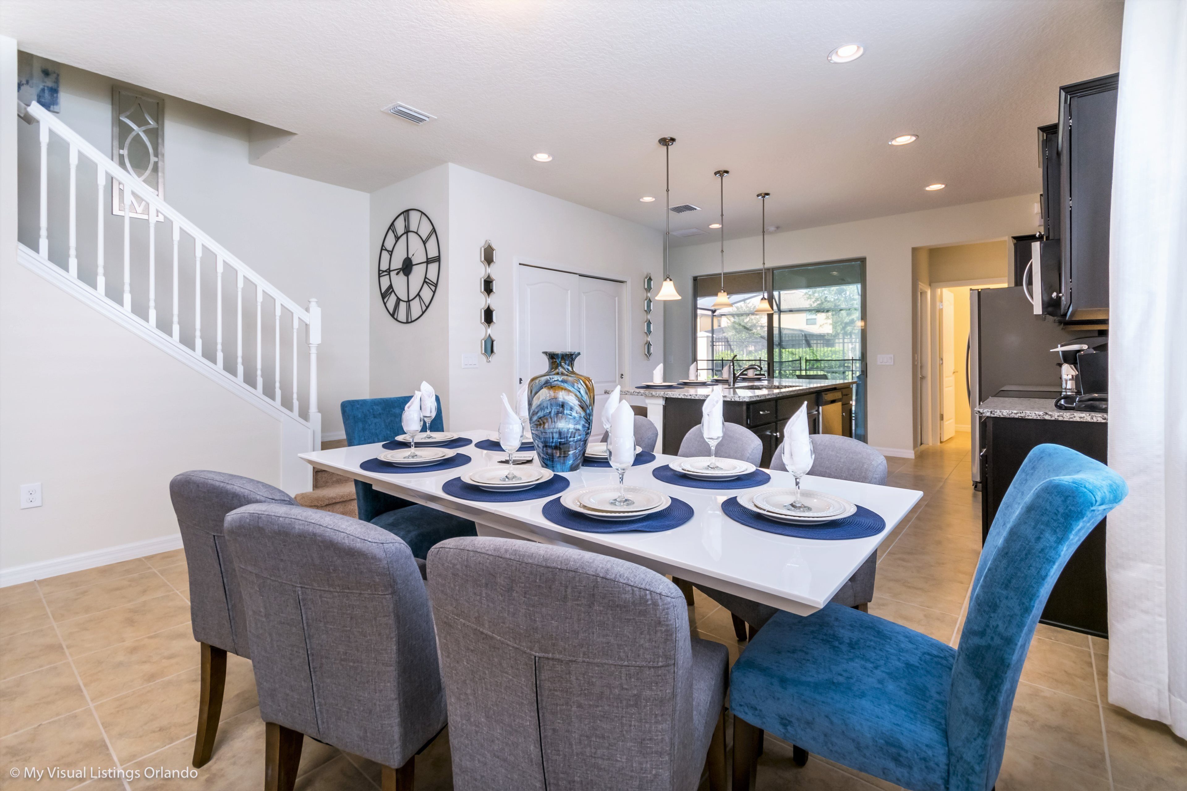A dining area with a white table set for six, grey and blue upholstered chairs, a large vase, and a clock on the wall.
