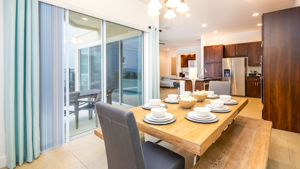A dining area with a wooden table set for four, a sliding glass door leading to a balcony, and a kitchen in the background. - 7770ll Ultimate Reunion Home (Kissimmee)
