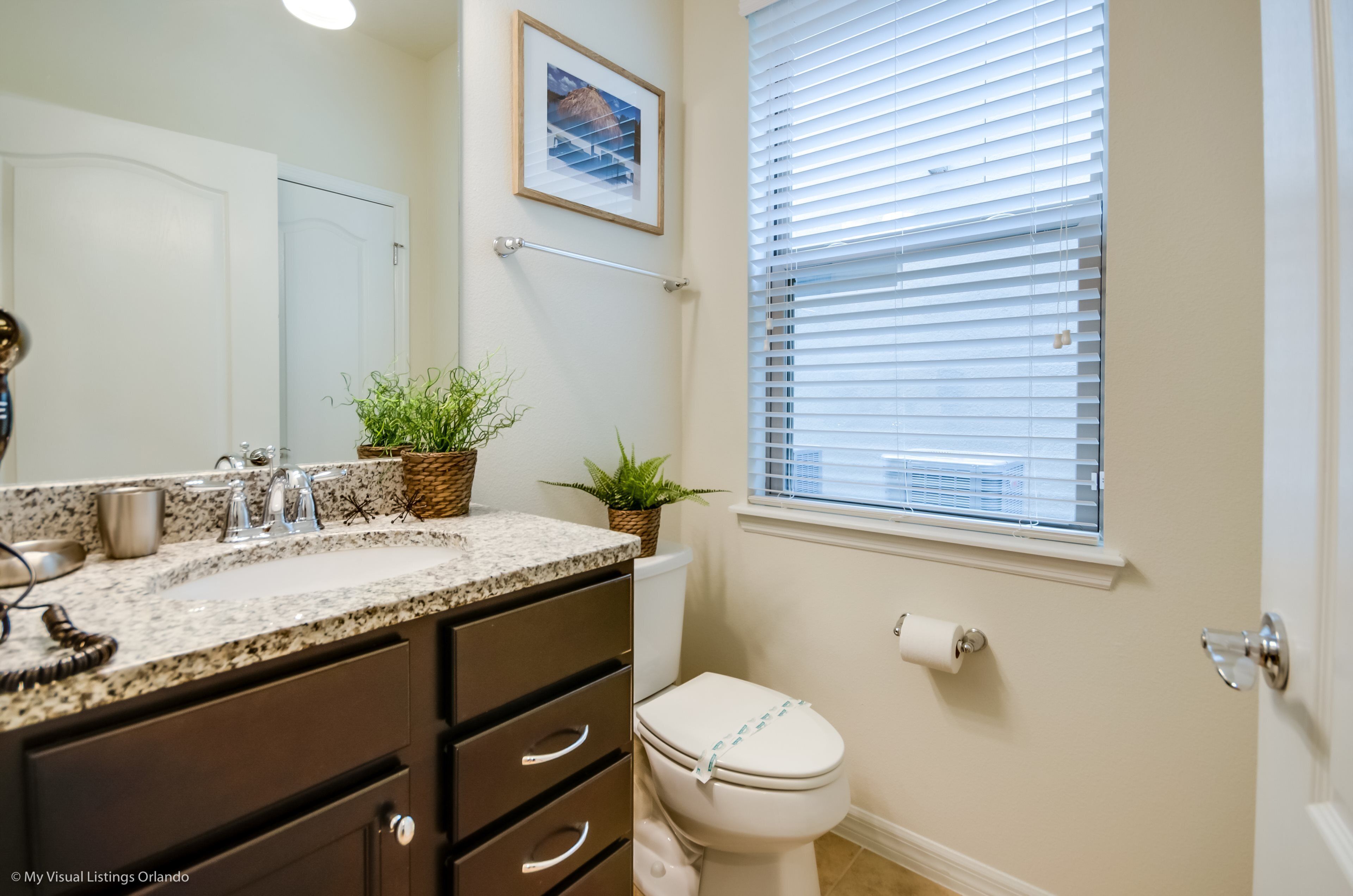 A bathroom with a white toilet, a sink with a granite countertop, a towel rack, and a window with blinds.