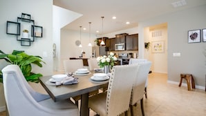 A dining area with a table set for four, surrounded by chairs, a kitchen in the background, and a living area with a sofa and coffee table. - Elegant & Fun 6 Bed Villa Games Pool Ihr4126 (Kissimmee)