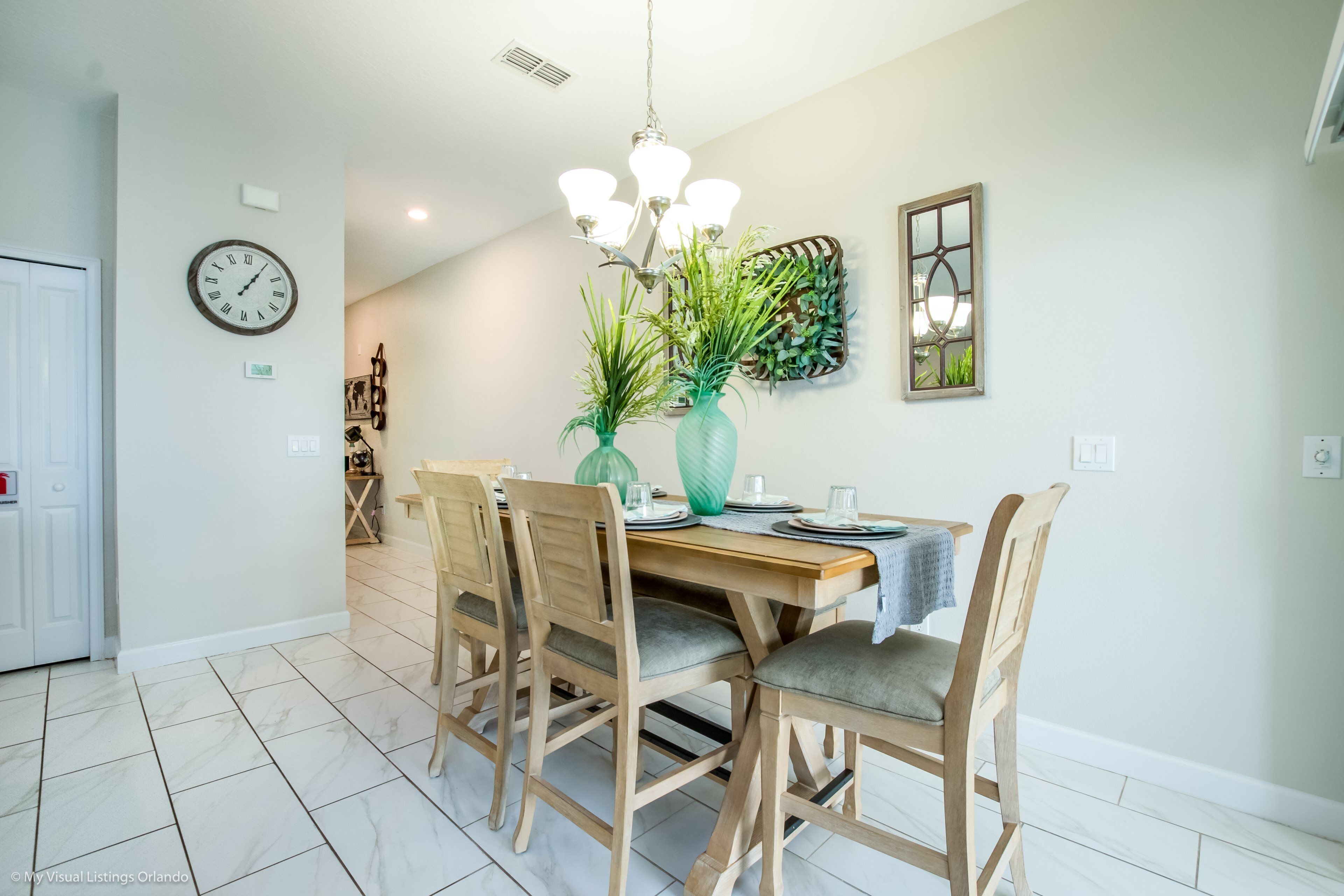 A dining area with a wooden table, four chairs, a green vase with plants, a wall clock, and a decorative mirror.