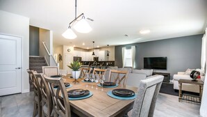 A dining area with a wooden table set for six, a kitchen in the background, and a living room with a TV. - 9 Bedroom Rental for 18 Guests (Davenport)