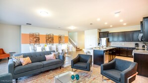 Ein modernes Wohnzimmer mit einem grauen Sofa, zwei Sesseln und einem Couchtisch. Eine Treppe führt zu einer Küche mit dunklen Schränken und Edelstahlgeräten.