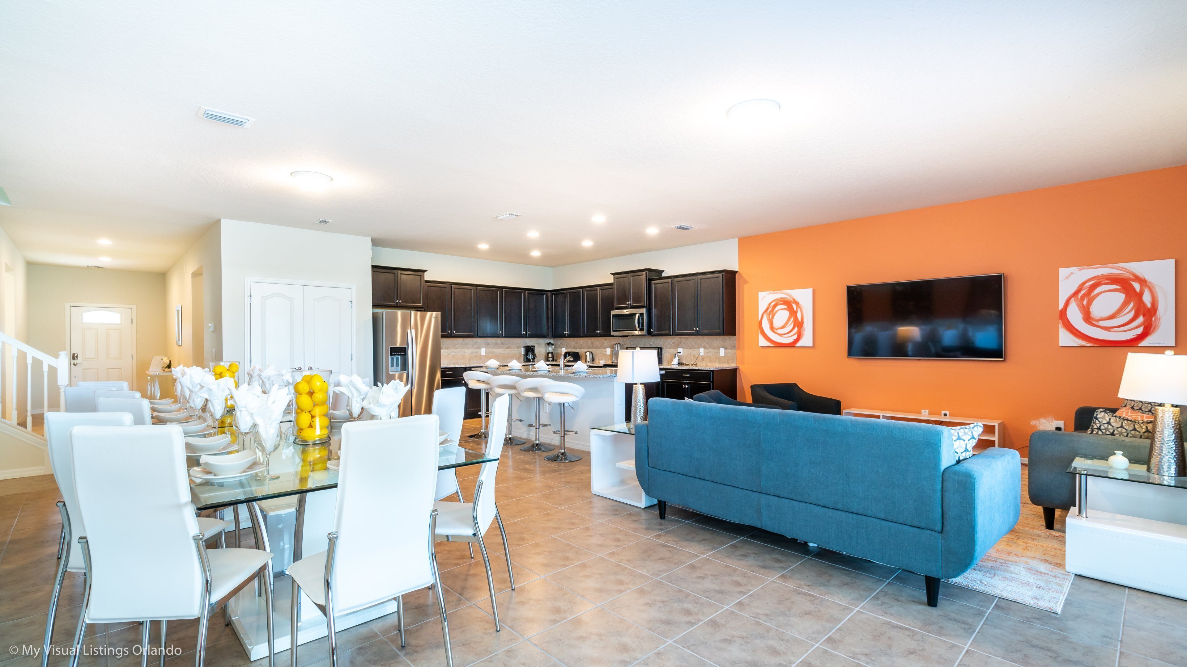 A modern living room with a blue sofa, white dining chairs, a glass table, and a kitchen in the background.