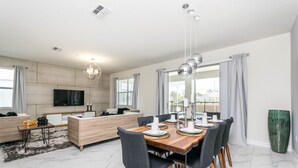 A modern living room with a wooden dining table set for four, a sofa, a TV mounted on the wall, and a large window with curtains. - 8 Bedroom Champions Gate Villa (Davenport)