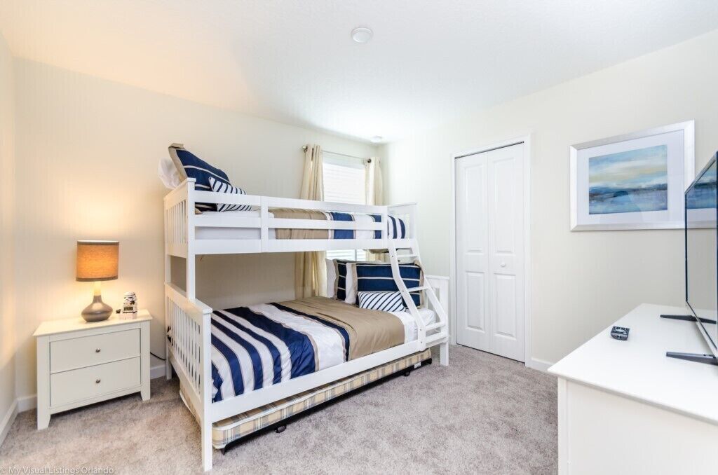 A bunk bed with striped bedding, a bedside table with a lamp, a desk with a computer, a door, and a framed picture on the wall.