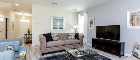 Una sala de estar con un sofá color beige, un sillón azul, una mesa de centro de vidrio y un televisor de pantalla plana sobre un soporte de madera oscura.