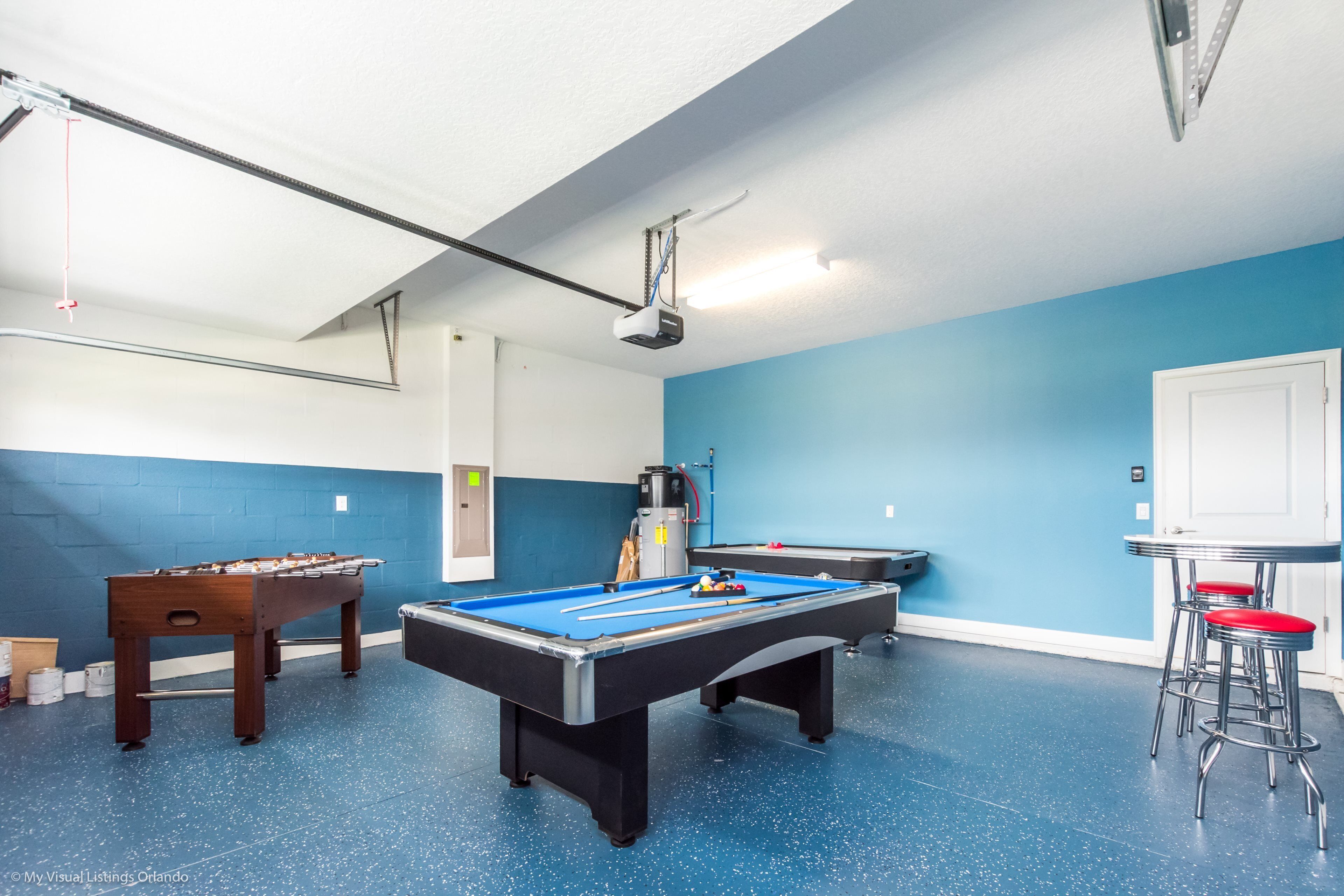 A pool table with a blue felt surface, a foosball table, and a pool table with a black felt surface. There are two bar stools with red seats.