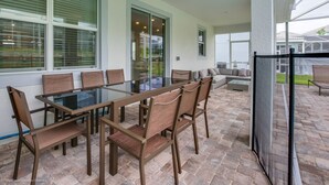 Een overdekt terras met een eettafel en stoelen met glazen blad, een bank en een glazen schuifdeur naar een achtertuin.