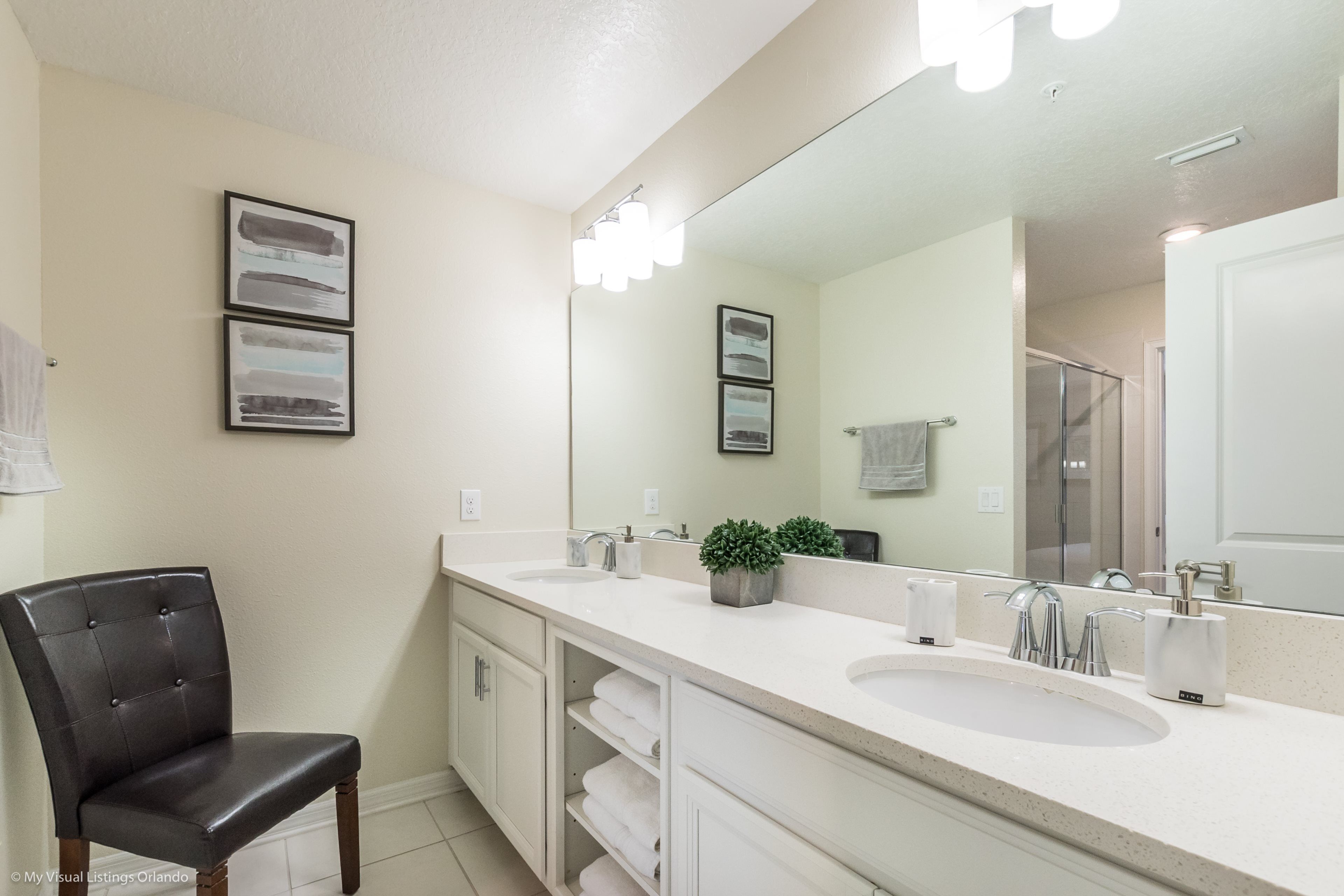 A modern bathroom with a large mirror, two sinks, a chair, and a potted plant.