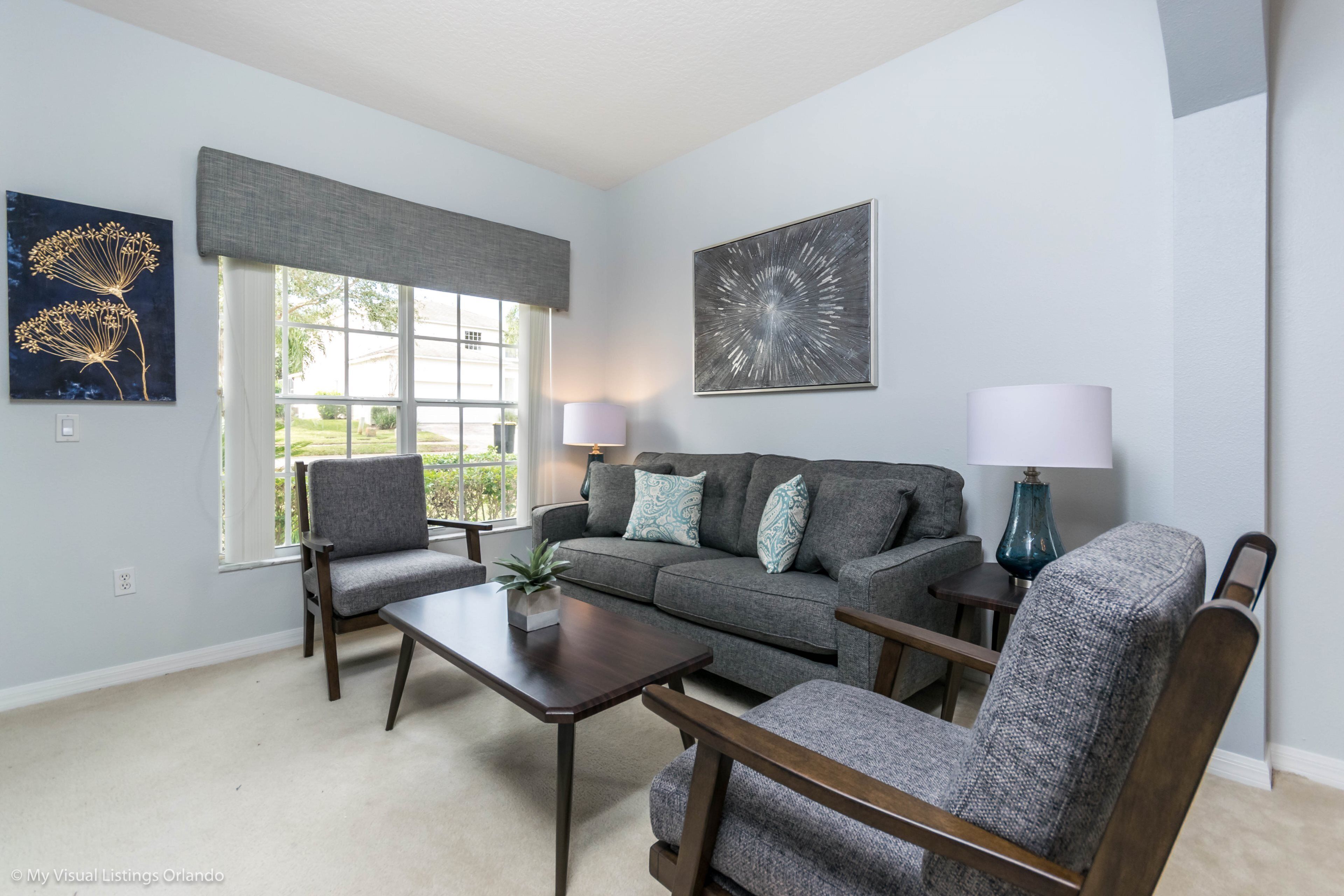 A living room with a grey sofa, two armchairs, a coffee table, and two lamps. There are two windows with curtains, and artwork on the walls.