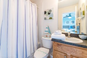 Ein Badezimmer mit gestreiftem Duschvorhang, einer Toilette mit dekorativem Segelboot, einem Waschbecken mit Handtuch und einem Spiegel mit Handtuchhalter.