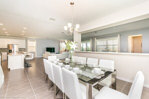 A spacious dining area with a glass table set for six, surrounded by white chairs. A modern chandelier hangs above, and the room features a large window with blinds. - Fabulous 9 Bed 6,5 Ba Villa Games, Pool & Spa 9063srl (Kissimmee)