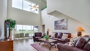 A spacious living room with a high ceiling, a large window, and a view of the outdoors. There is a brown leather sofa set, a coffee table, and a TV on a wooden stand. - 526wp 5 bed 4 Bathroom Overlooking Amazing View (Davenport)