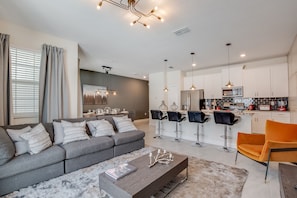 A modern living room with a grey sofa, a coffee table, and a kitchen area with white cabinets and a bar. - 4 Bedroom Town Home with Pool (Kissimmee)