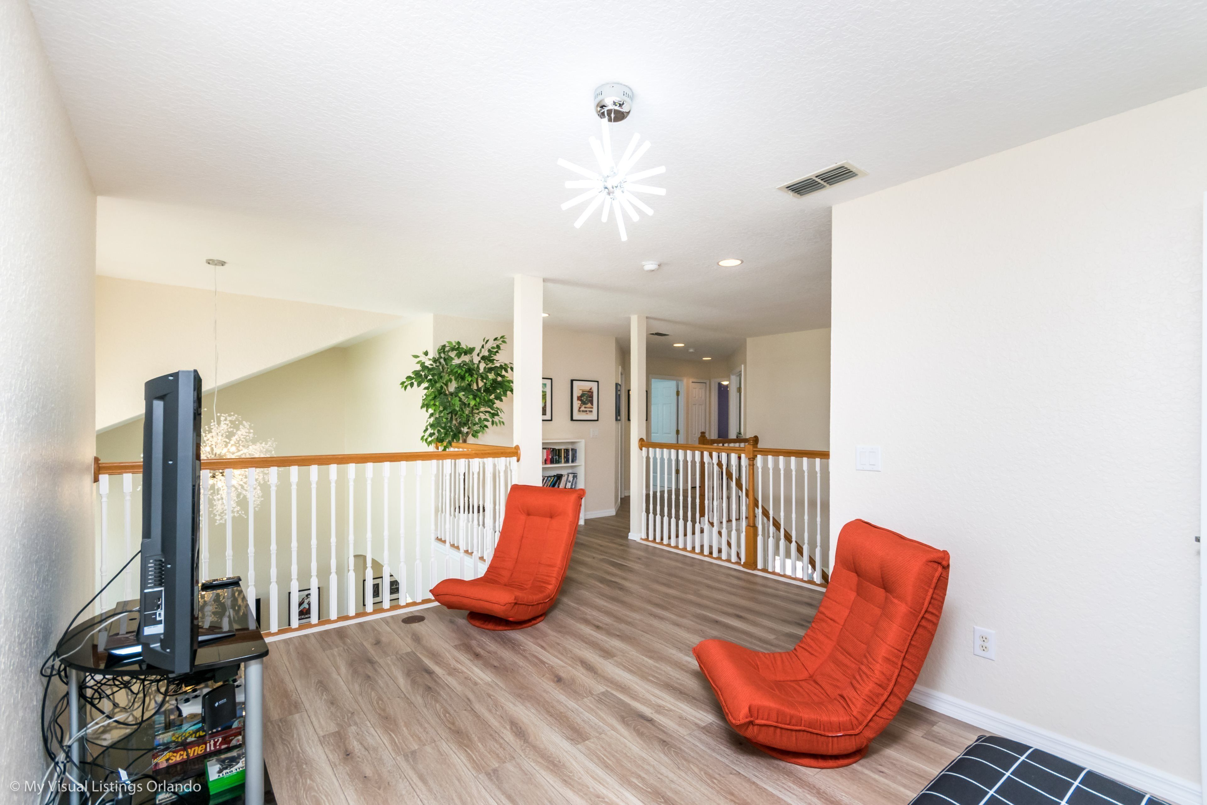 A spacious living area with a wooden floor, two orange chairs, a television on a stand, and a staircase leading to another level.