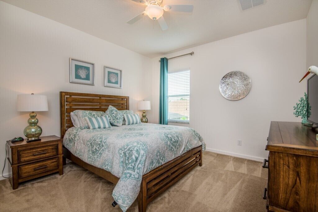 A bedroom with a wooden bed, two bedside tables with lamps, a ceiling fan, and a wall-mounted TV.